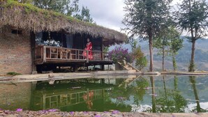 A traditional thatched-roof hut with a wooden balcony, a small pond in front, and a mountainous backdrop. - Hello Mu Cang Chai Homestay - Hostel (Pung Luong)