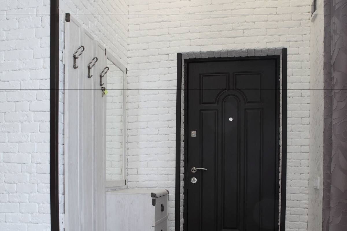 Um corredor com parede de tijolos brancos, porta preta e um banco com assento claro.