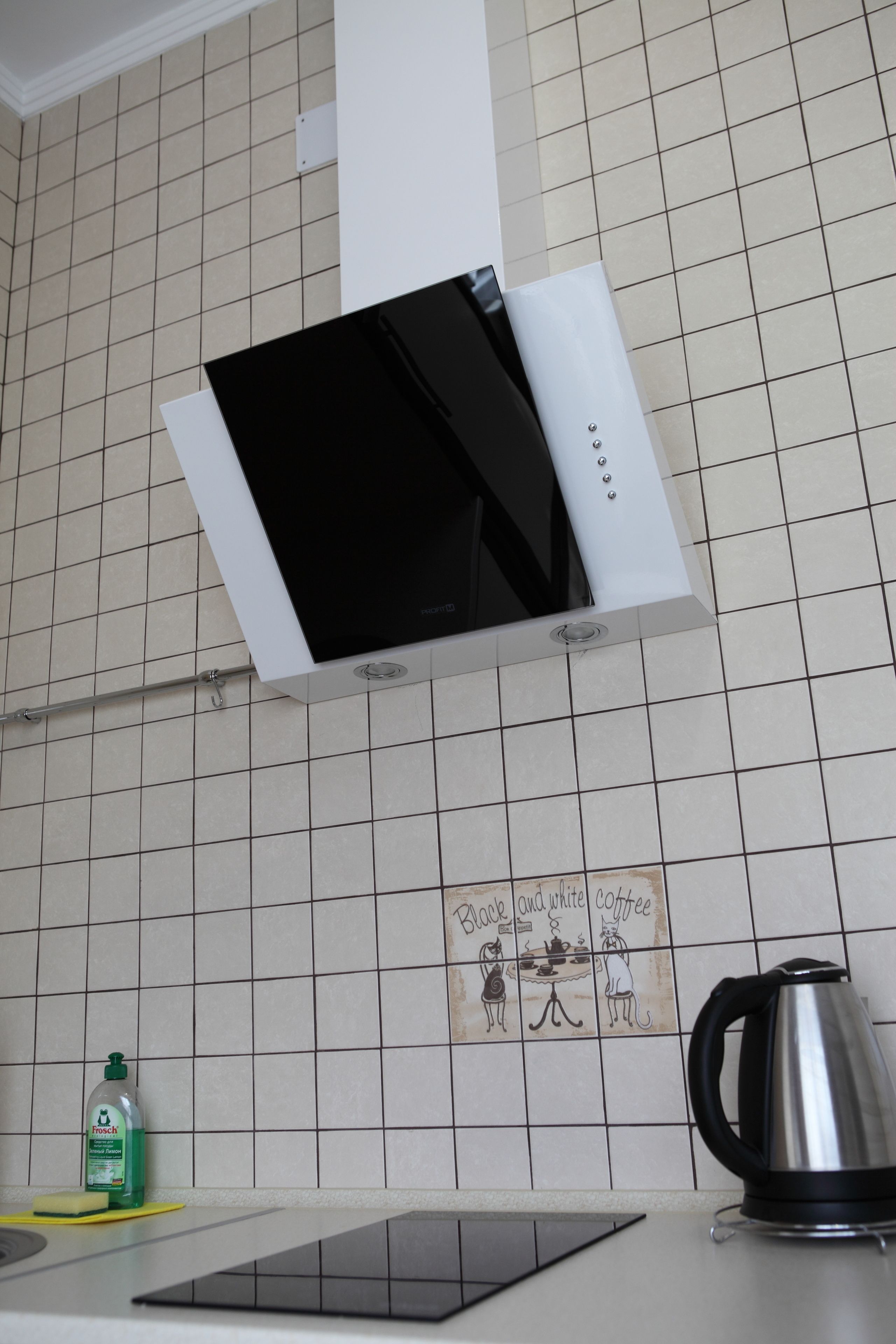 A kitchen with a white tiled backsplash, a black and white coffee-themed tile, a stainless steel kettle, and a bottle of dish soap.