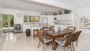 A spacious kitchen with white cabinetry, a dining area with wicker chairs, and a large window offering a view of greenery. - Natura Beach Pool Villa 2 (Rarotonga)