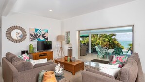 A modern living room with a sofa, coffee table, and artwork on the wall. There is a television and a lamp on a wooden stand. - Natura Beach Pool Villa 2 (Rarotonga)