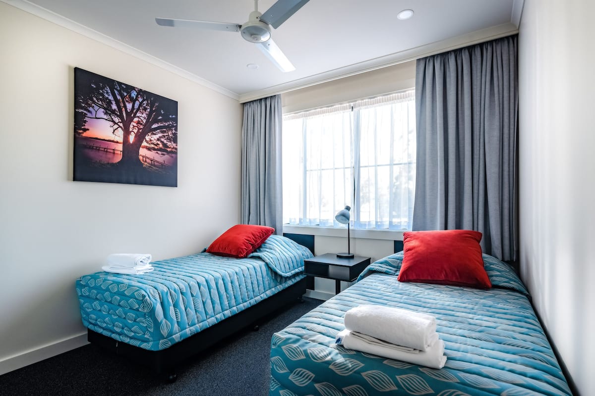 Habitación de hotel con dos camas, cada una con una colcha estampada azul y almohadas rojas. En la pared, hay un cuadro de un atardecer, un ventilador de techo y una ventana con cortinas.