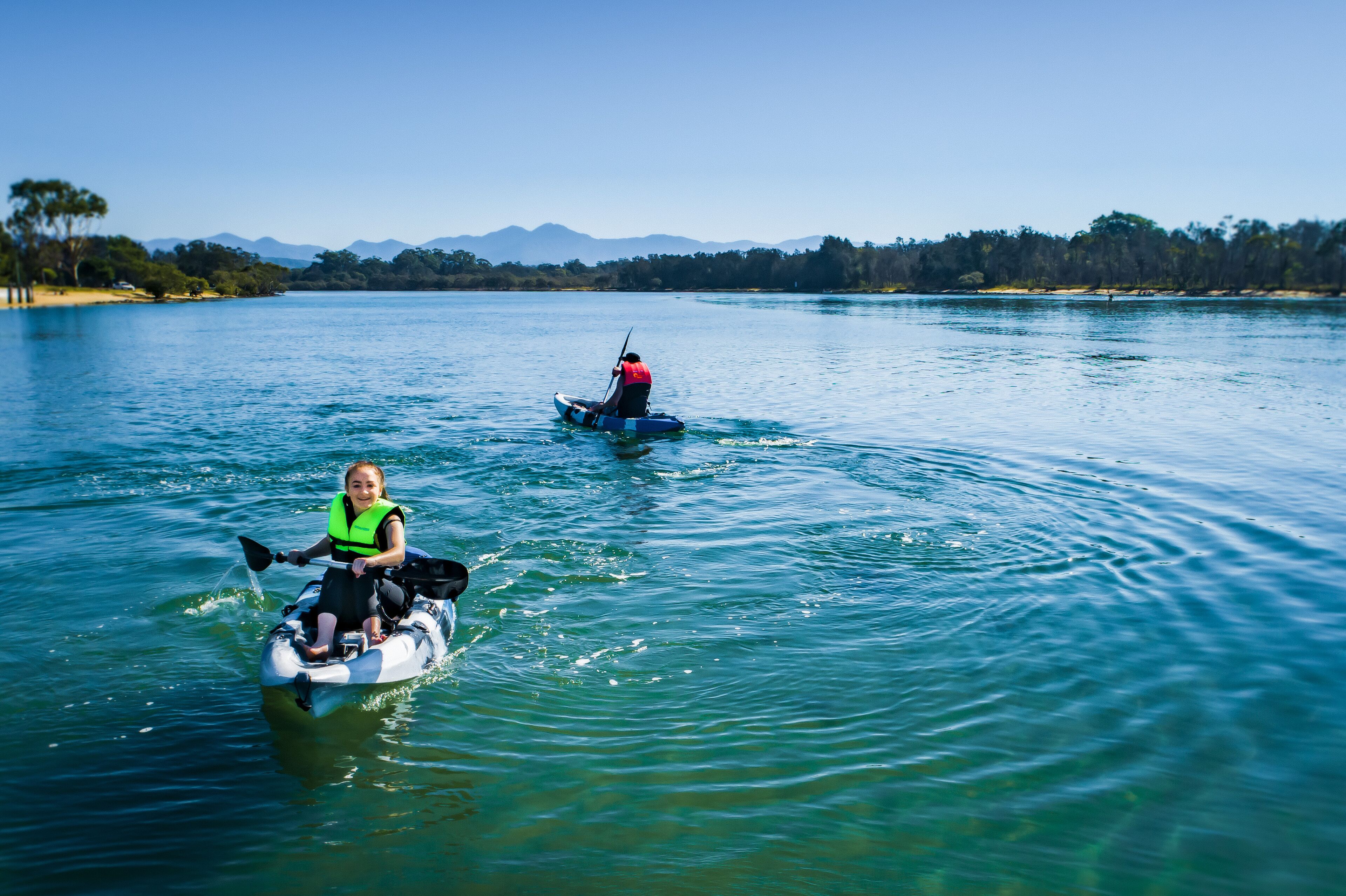 Deux personnes à bord d’un embarcation sur un lac paisible, avec des arbres et des montagnes en arrière-plan.