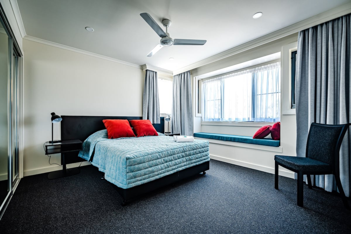 Una habitación de hotel moderna con cama, escritorio, silla, ventilador de techo y ventana con cortinas.