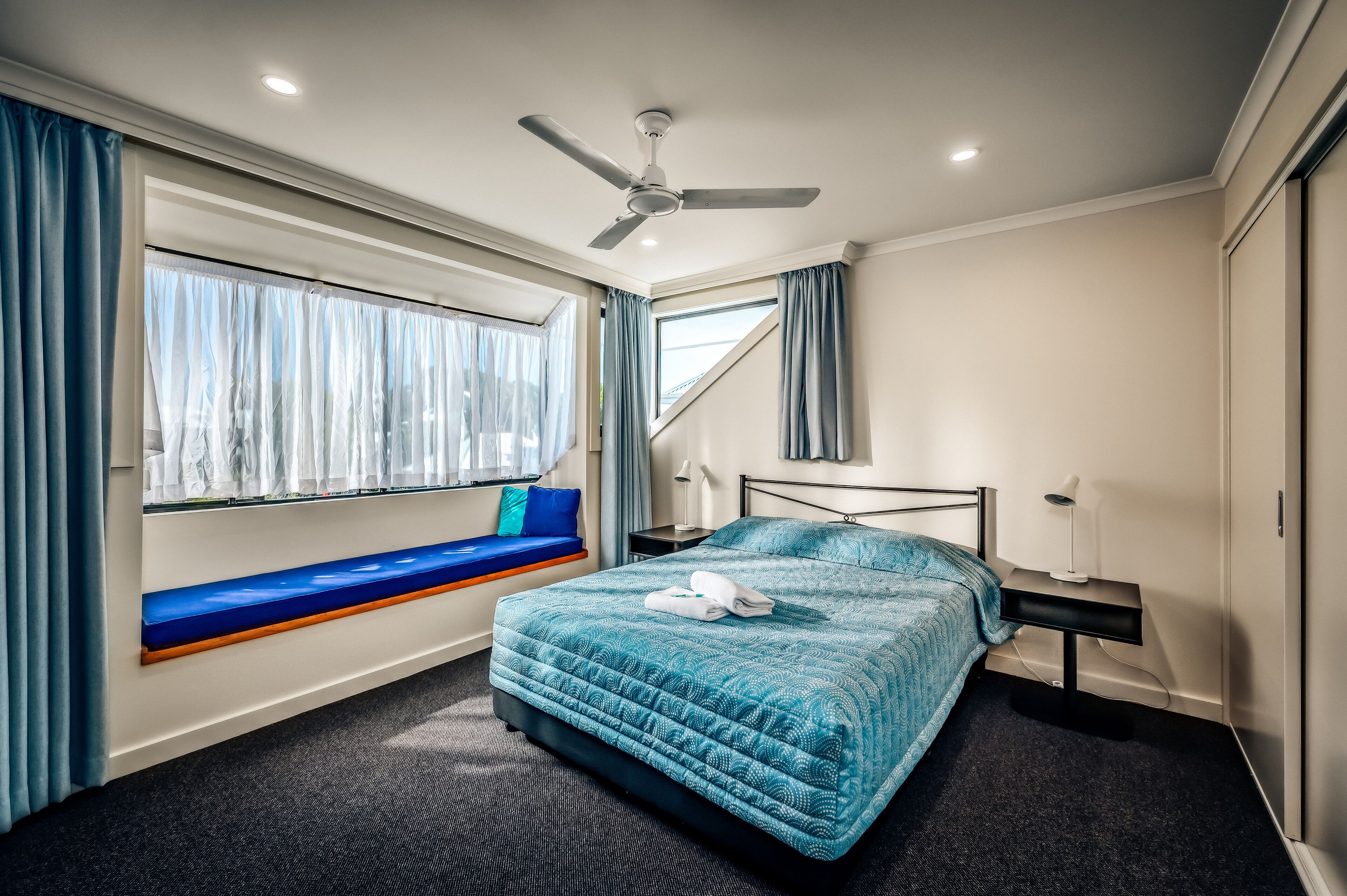 A bedroom with a bed, a window seat, a ceiling fan, and a nightstand.