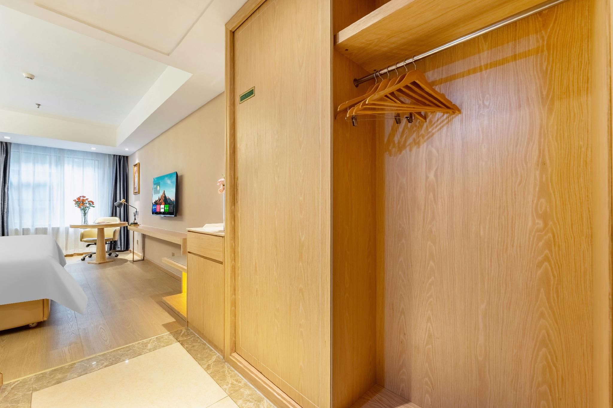 A hotel room with a wooden wardrobe, a desk with a chair, a TV on the wall, and a view of the room.