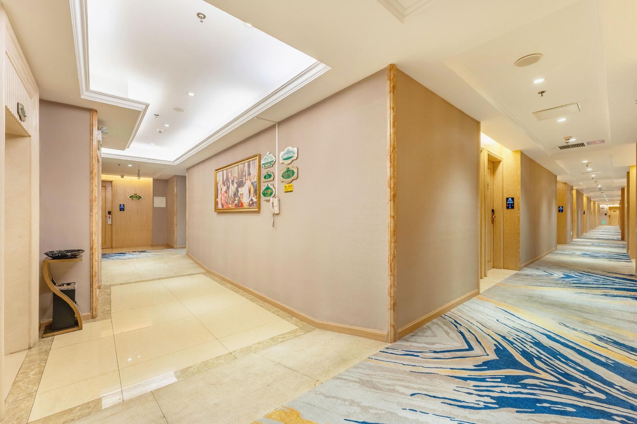 A hotel corridor with a patterned carpet, a framed artwork on the wall, and a trash bin.