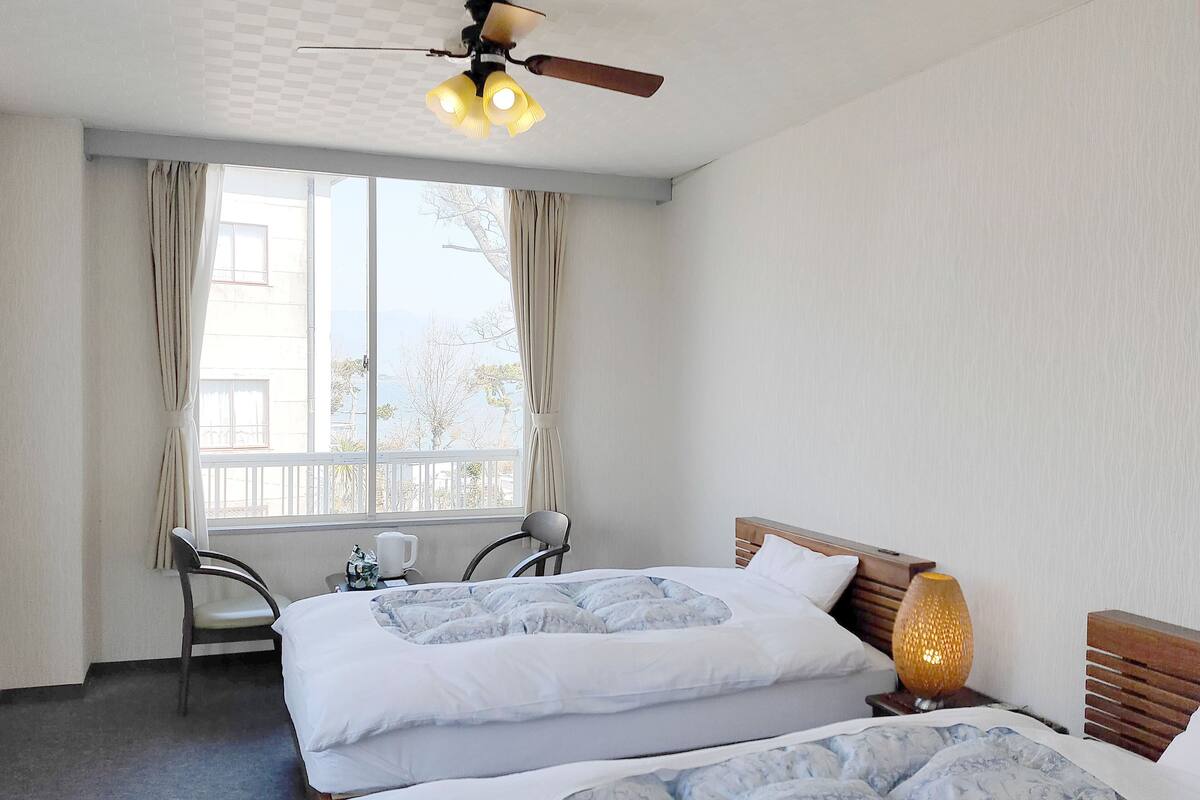 A hotel room with two beds, a ceiling fan, a window with curtains, a chair, and a bedside table with a lamp.