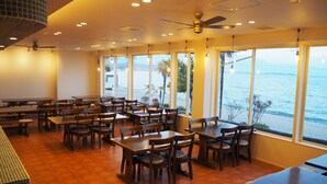 A restaurant with tables and chairs arranged in rows, large windows with a view of the sea, and a ceiling fan. - View Lodge Biwa (Otsu)