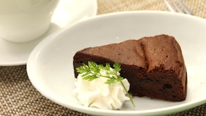 A slice of chocolate cake on a white plate with a dollop of whipped cream and a sprig of parsley, accompanied by a white cup and saucer. - Yamanakako Garden Villa (Yamanakako)
