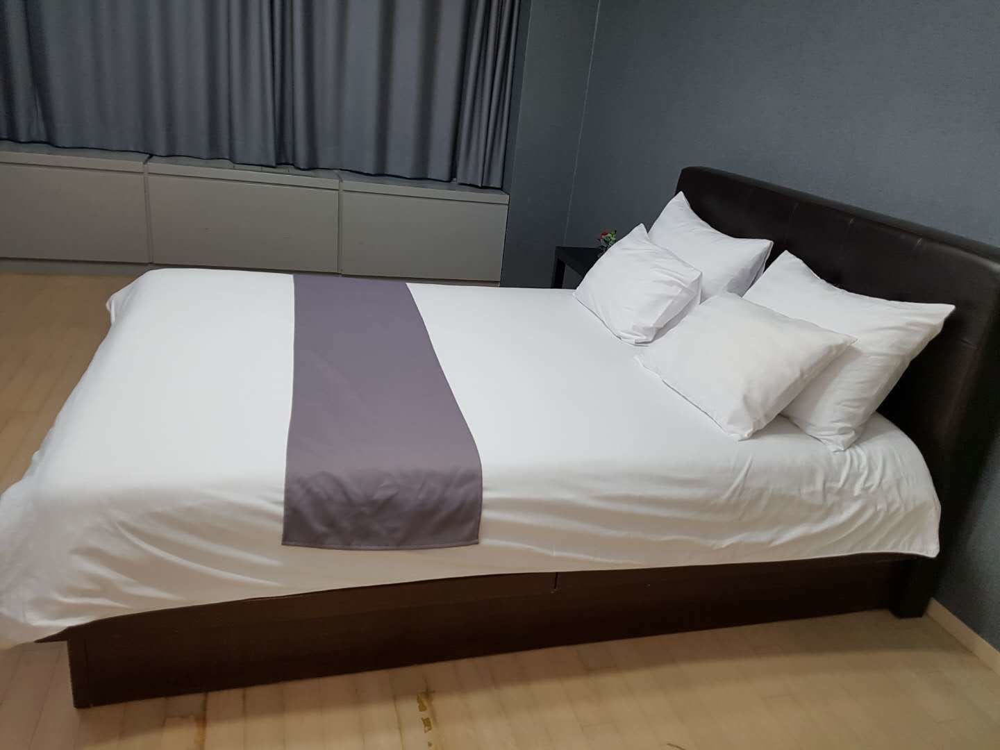 Una cama bien hecha con ropa de cama blanca y morada, un cabecero oscuro y suelo de madera.