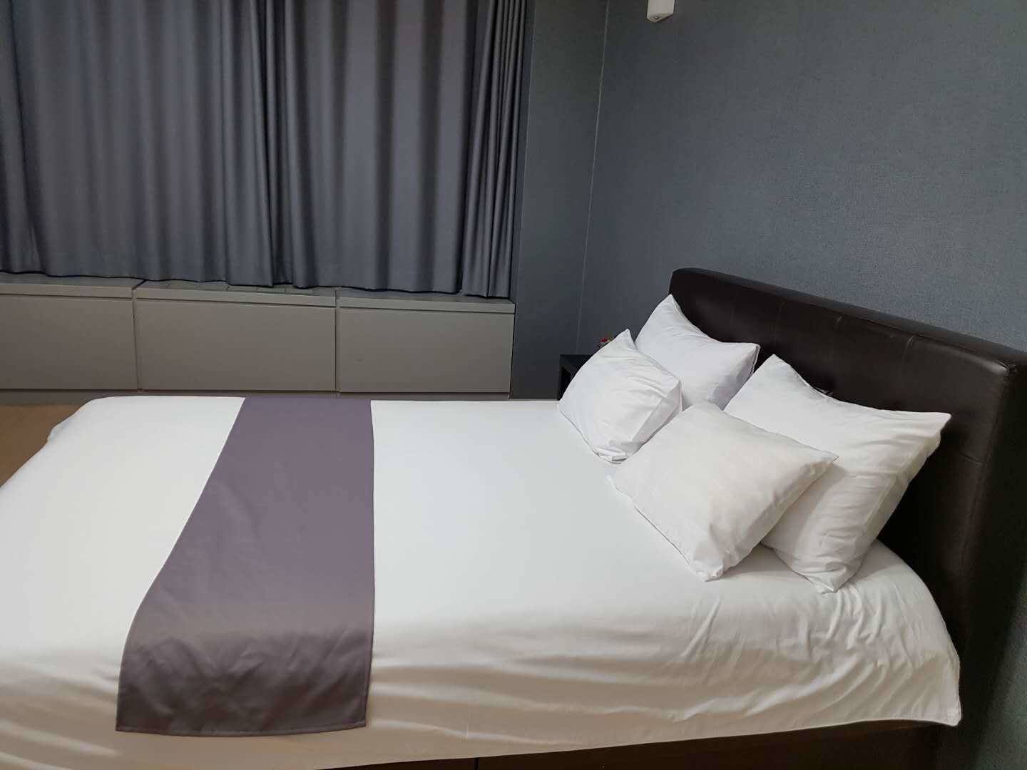 A neatly made bed with white and gray linens and two pillows, a gray accent pillow, and a dark headboard in a room with gray walls and a window with curtains.