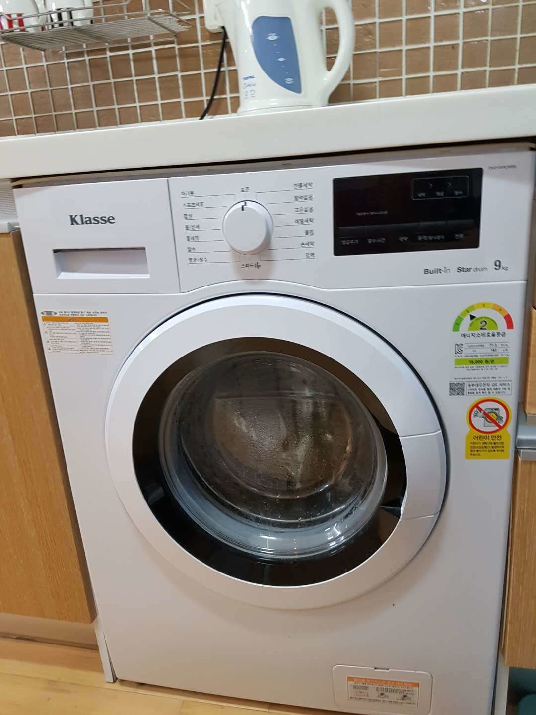 A front-loading washing machine with a control panel, a built-in steam function, and a safety warning sticker.