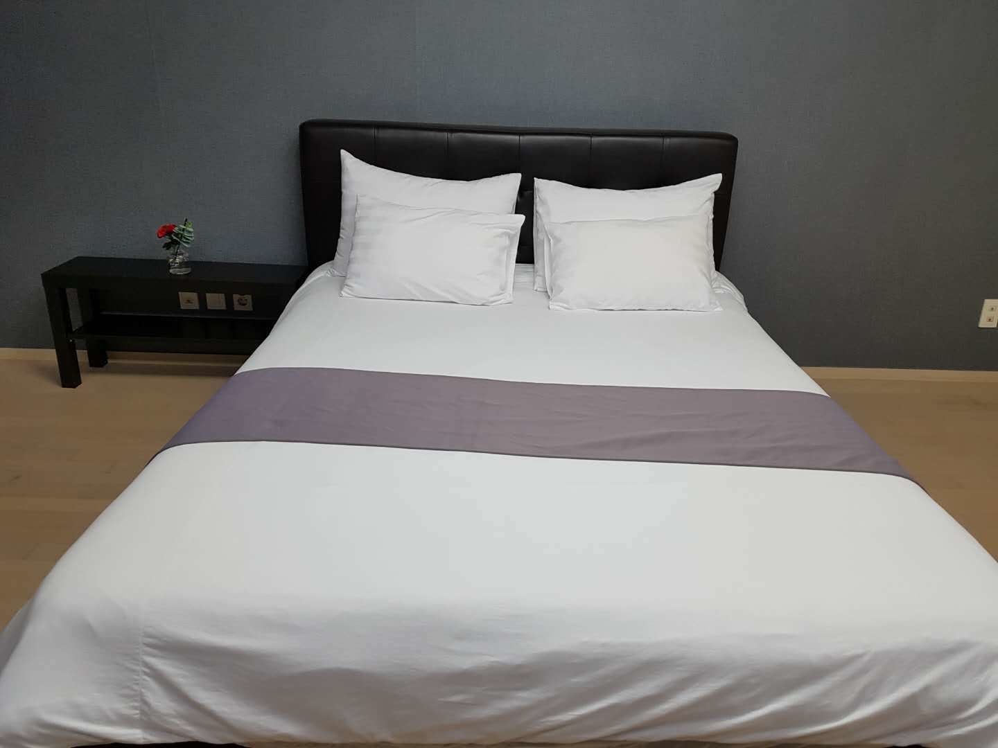 A neatly made bed with white bedding and a grey accent, a black headboard, and a small bedside table with a red flower.