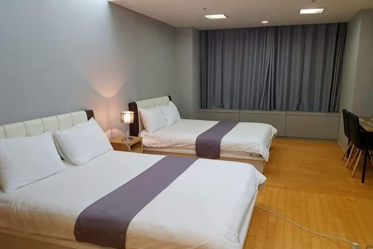 A hotel room with two beds, a desk, a chair, a window with curtains, and a lamp.
