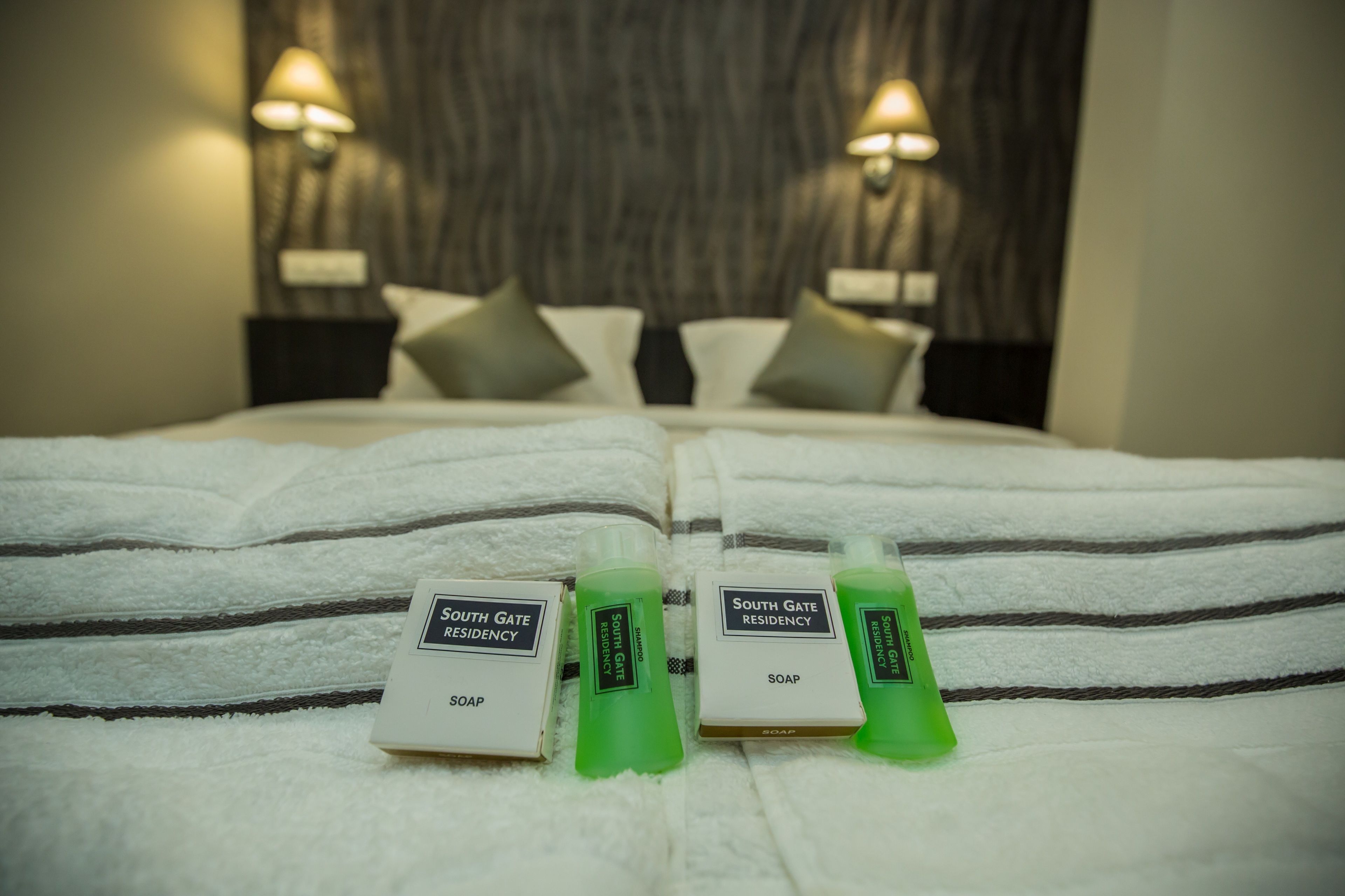 Una cama bien hecha con dos botellas verdes de jabón del South Gate Residency y una almohada.