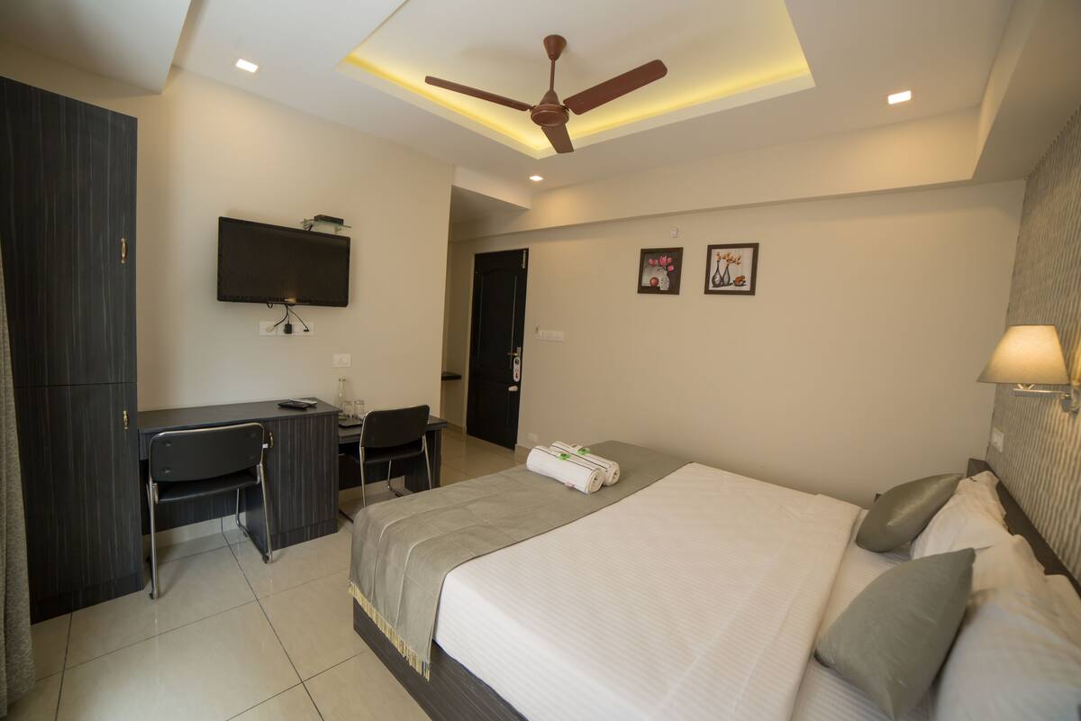 A modern hotel room with a bed, desk, chair, ceiling fan, TV, and artwork on the wall.