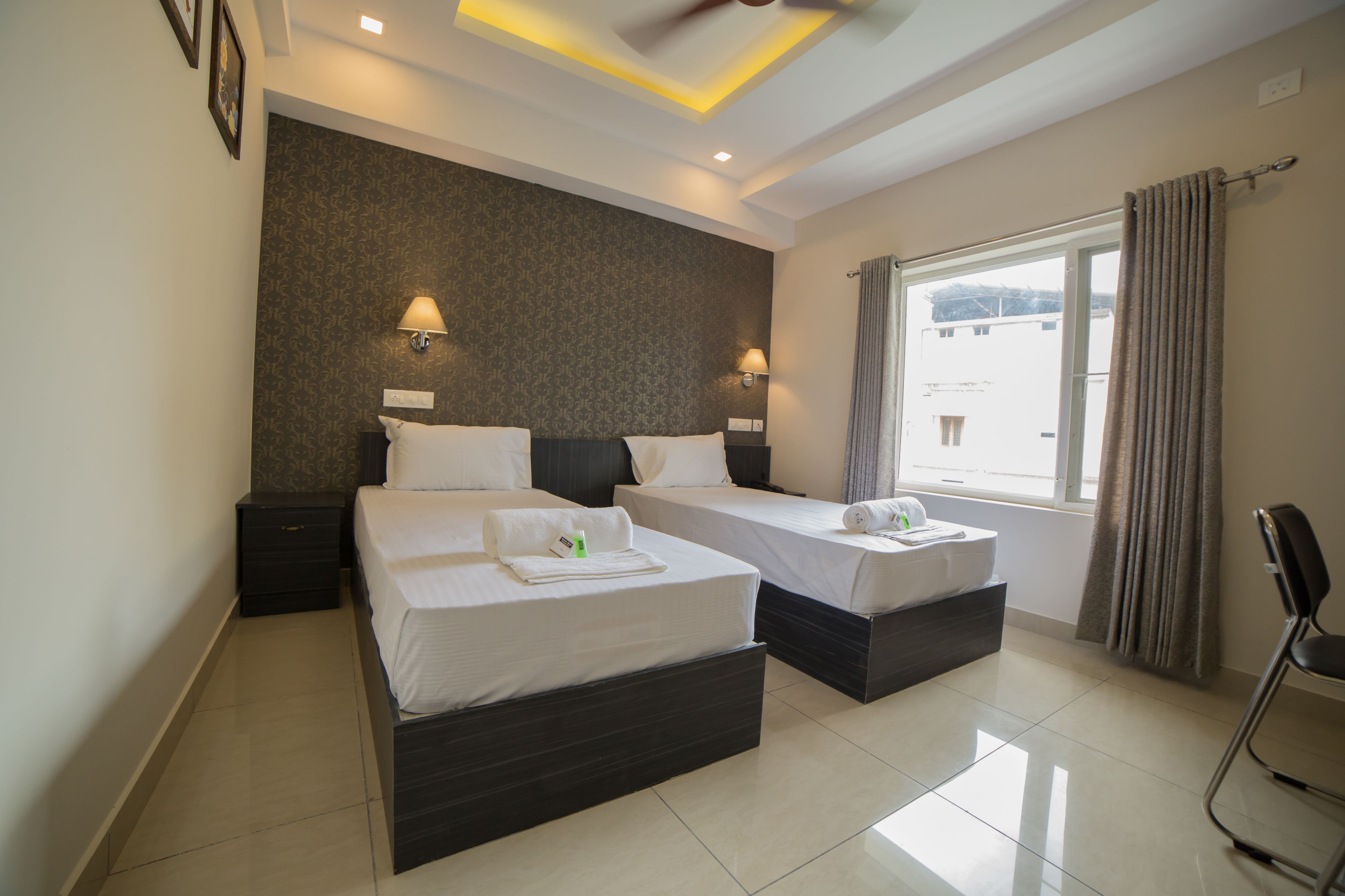 A hotel room with two beds, a window with curtains, a chair, and a wall-mounted lamp.