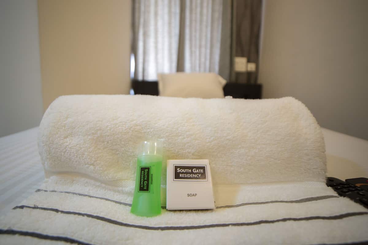 A hotel bed with a white towel, a green bottle of South Gate Residency soap, and a remote control.