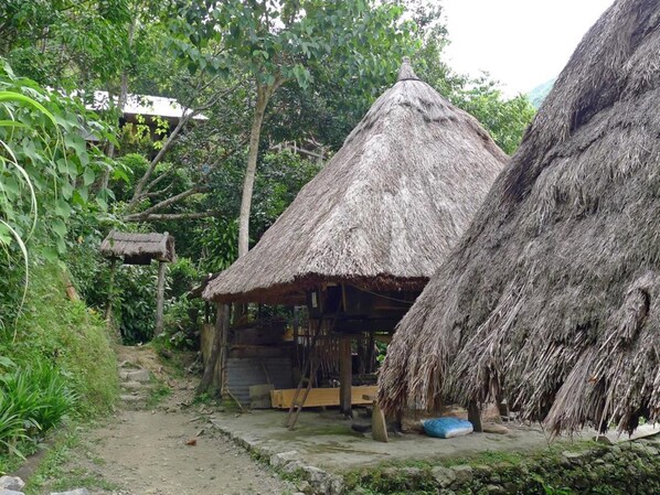 Uma cabana rústica com telhado de palha, um banco de madeira e uma pequena mesa, rodeada por vegetação exuberante.