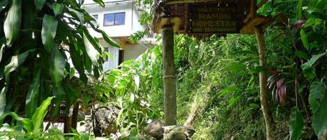 A rustic thatched-roof sign welcoming guests to Ramon's Home Stay and Restaurant, surrounded by lush greenery and a stone wall.
