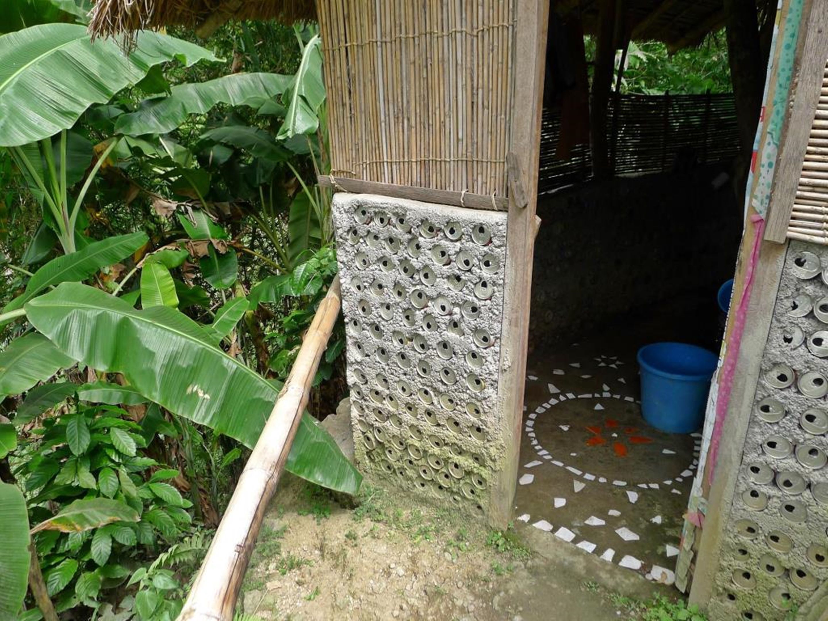 Une cabane rustique avec un toit en bambou, un mur à la texture particulière orné de trous circulaires et un sol carrelé avec un motif circulaire.