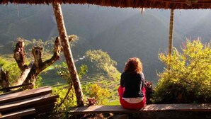 A person sitting on a wooden bench under a thatched roof, overlooking a terraced landscape. - Ramons Native Homestay and Restaurant (Banaue)