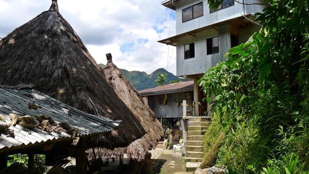 A rustic village with a thatched-roof hut, a corrugated metal structure, and a staircase leading to a two-story building.