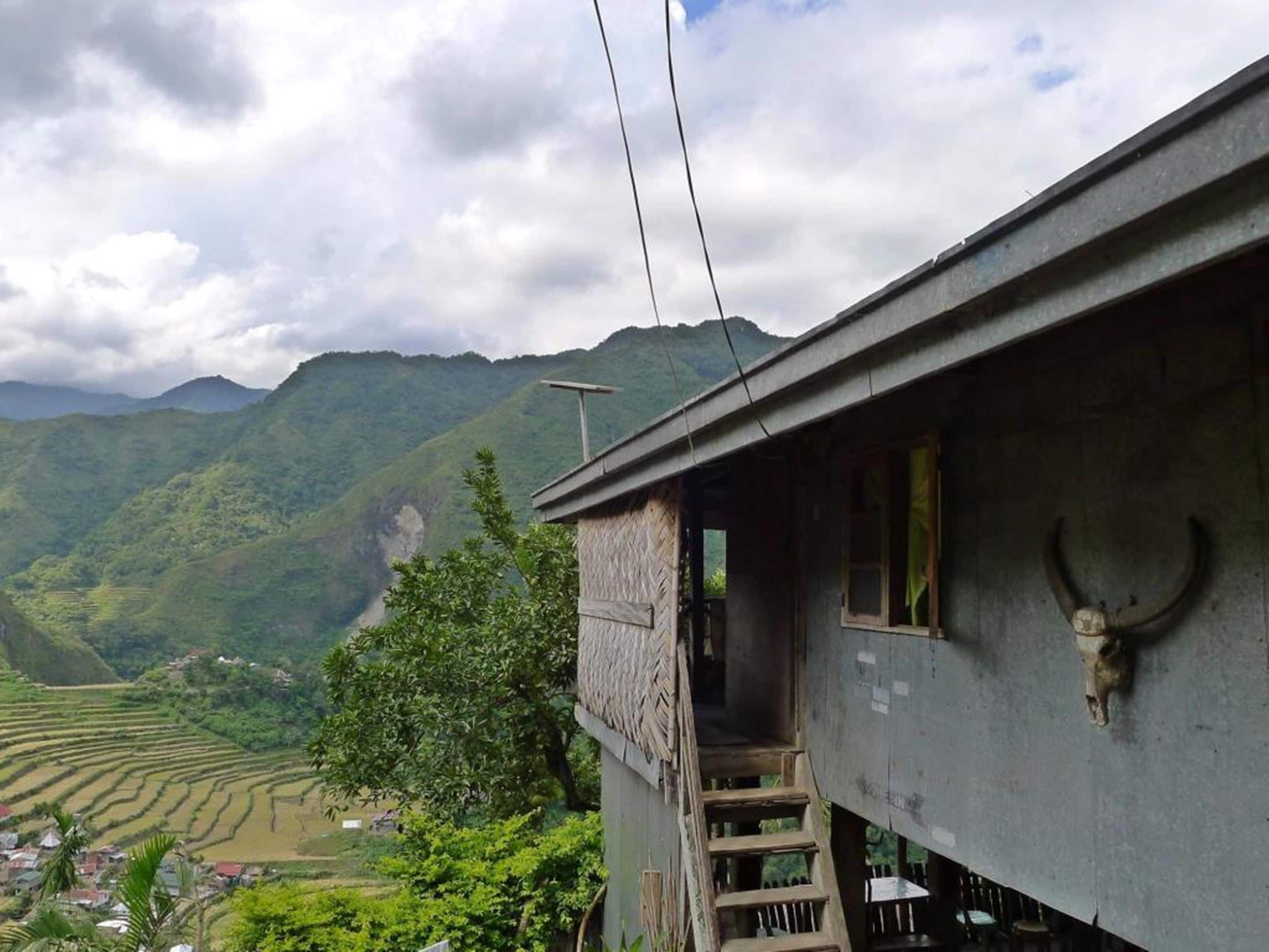A house with a staircase leading to a porch, surrounded by lush green hills and a cloudy sky.