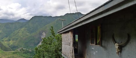 A house with a staircase leading to a porch, surrounded by lush green hills and a cloudy sky.