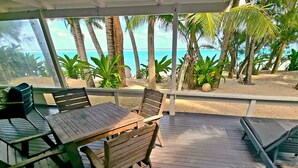 Un espace extérieur aménagé, abrité et doté de mobilier en bois, offrant une vue sur une plage bordée de palmiers et sur l’océan.