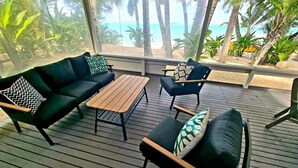 Un espace extérieur aménagé, abrité et doté d’une table en bois et de deux canapés, dont l’un est agrémenté d’un coussin à motifs et l’autre d’un coussin uni, et qui offre une vue sur une plage bordée de palmiers.