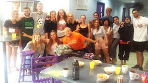 Un grupo de personas reunidas alrededor de una mesa con comida y bebidas, en una sala con un letrero que dice "FAITH FAMILY FRIENDS".