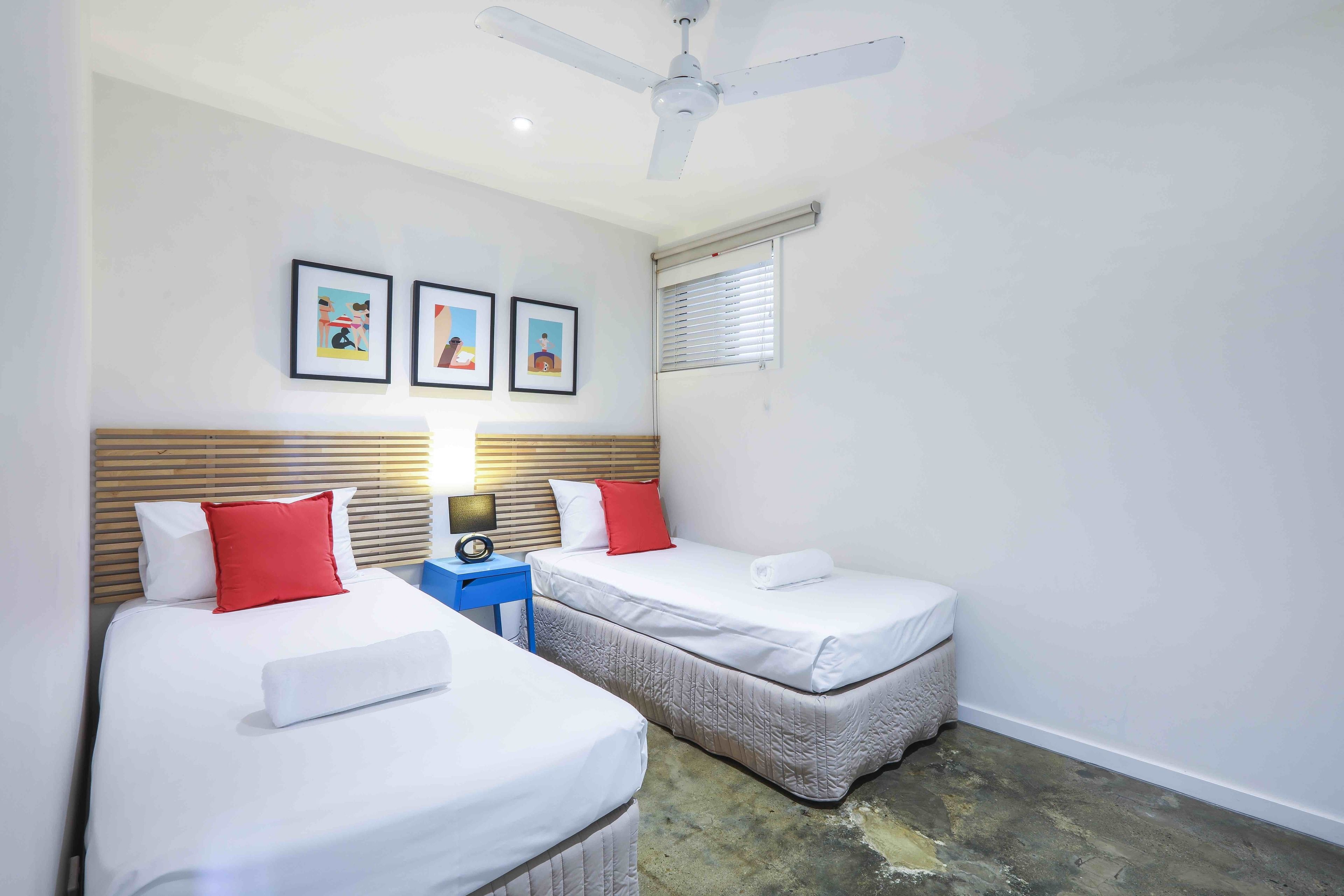 Une chambre d’hôtel avec deux lits, un ventilateur de plafond, une fenêtre avec des stores et trois tableaux encadrés accrochés au mur.