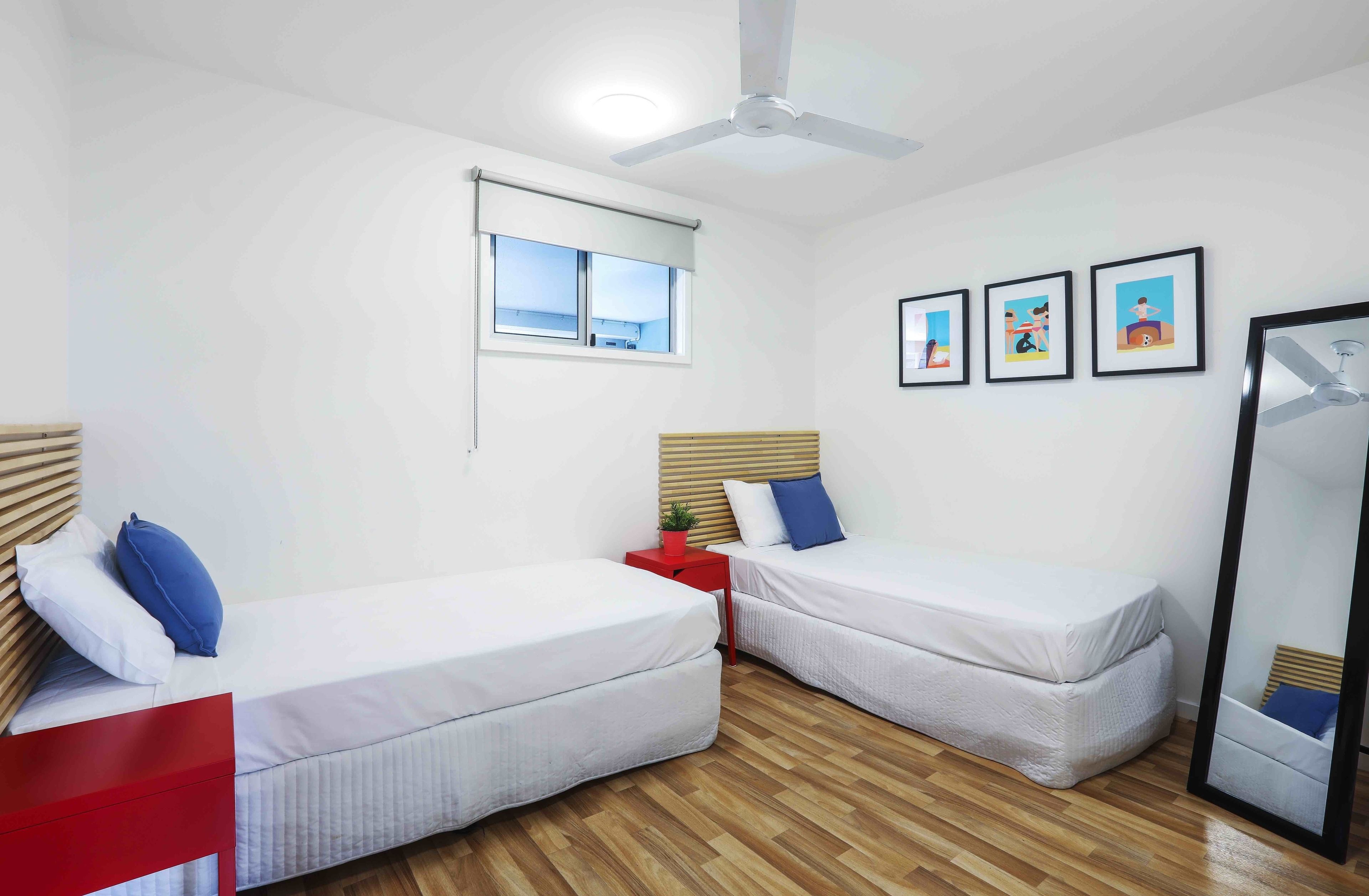Una habitación con dos camas individuales, un ventilador de techo, un ventanal con persianas y tres cuadros en la pared.