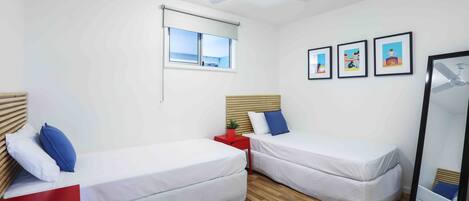 Una habitación con dos camas individuales, un ventilador de techo, un ventanal con persianas y tres cuadros en la pared.