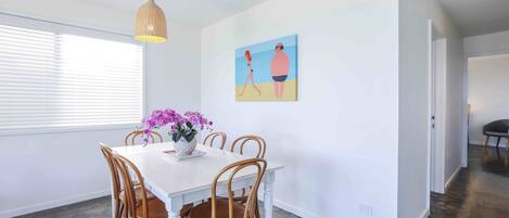 A dining area with a white table and wooden chairs, a hanging pendant light, and a painting on the wall.
