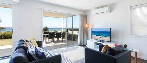 Ein modernes Wohnzimmer mit großem Fernseher, zwei Sofas, einem kleinen Tisch und Meerblick durch große Schiebetüren.