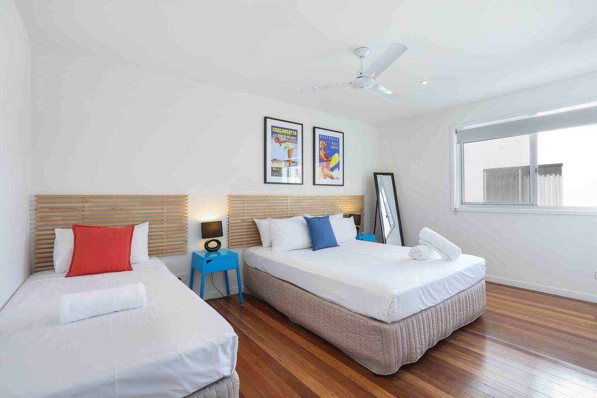 A hotel room with two beds, wooden flooring, a ceiling fan, and two framed posters on the wall.