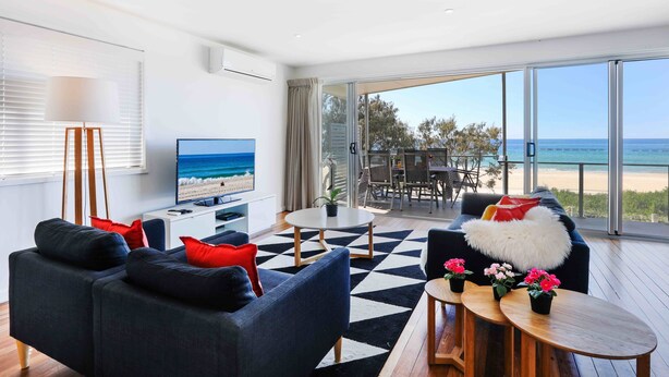 Ein modernes Wohnzimmer mit großem Fernseher, bequemen Sitzmöglichkeiten, einem Couchtisch und Blick auf den Strand durch große Fenster.
