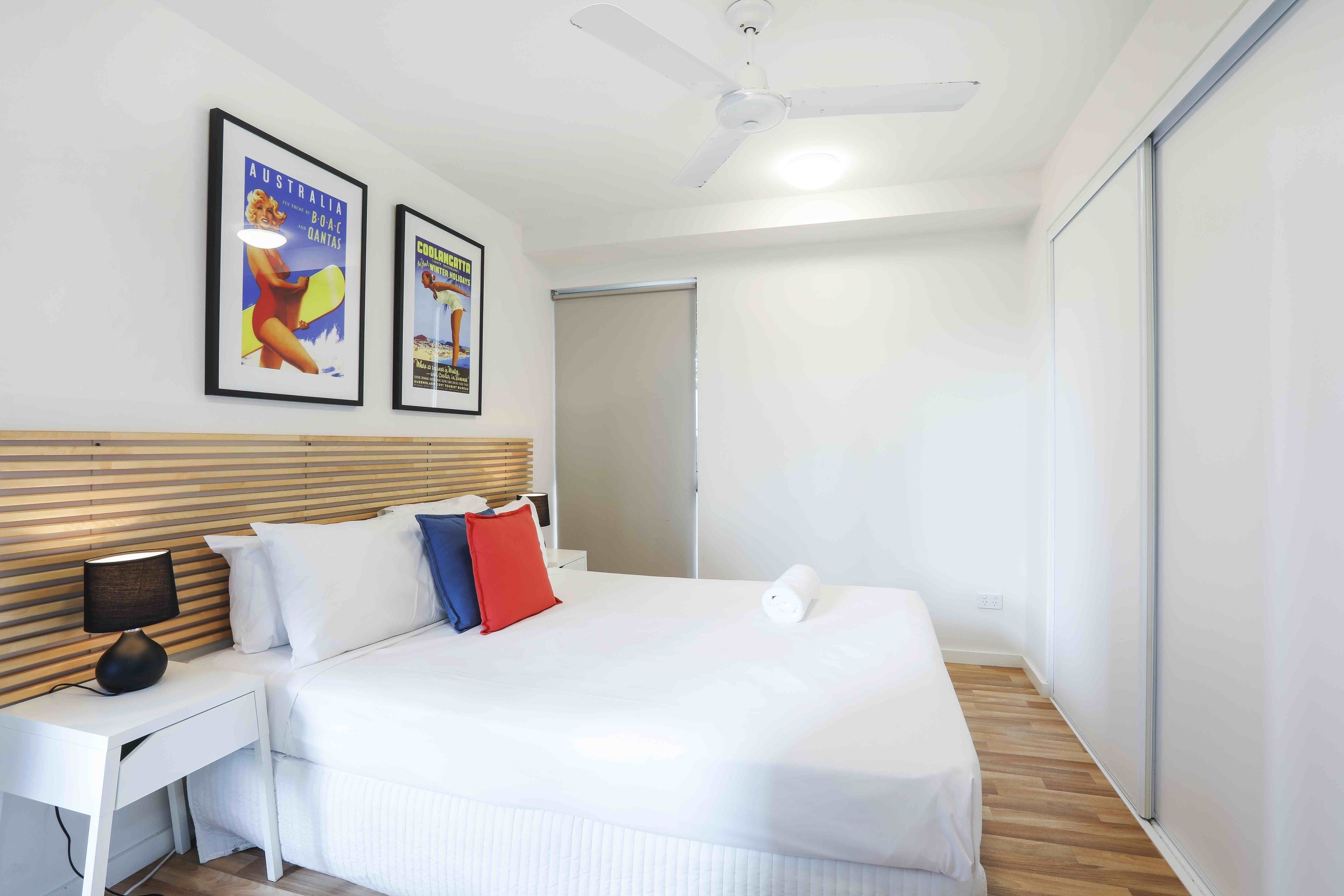 Quarto de hotel moderno com uma cama grande, uma mesinha de cabeceira com abajur, um ventilador de teto e dois quadros na parede.