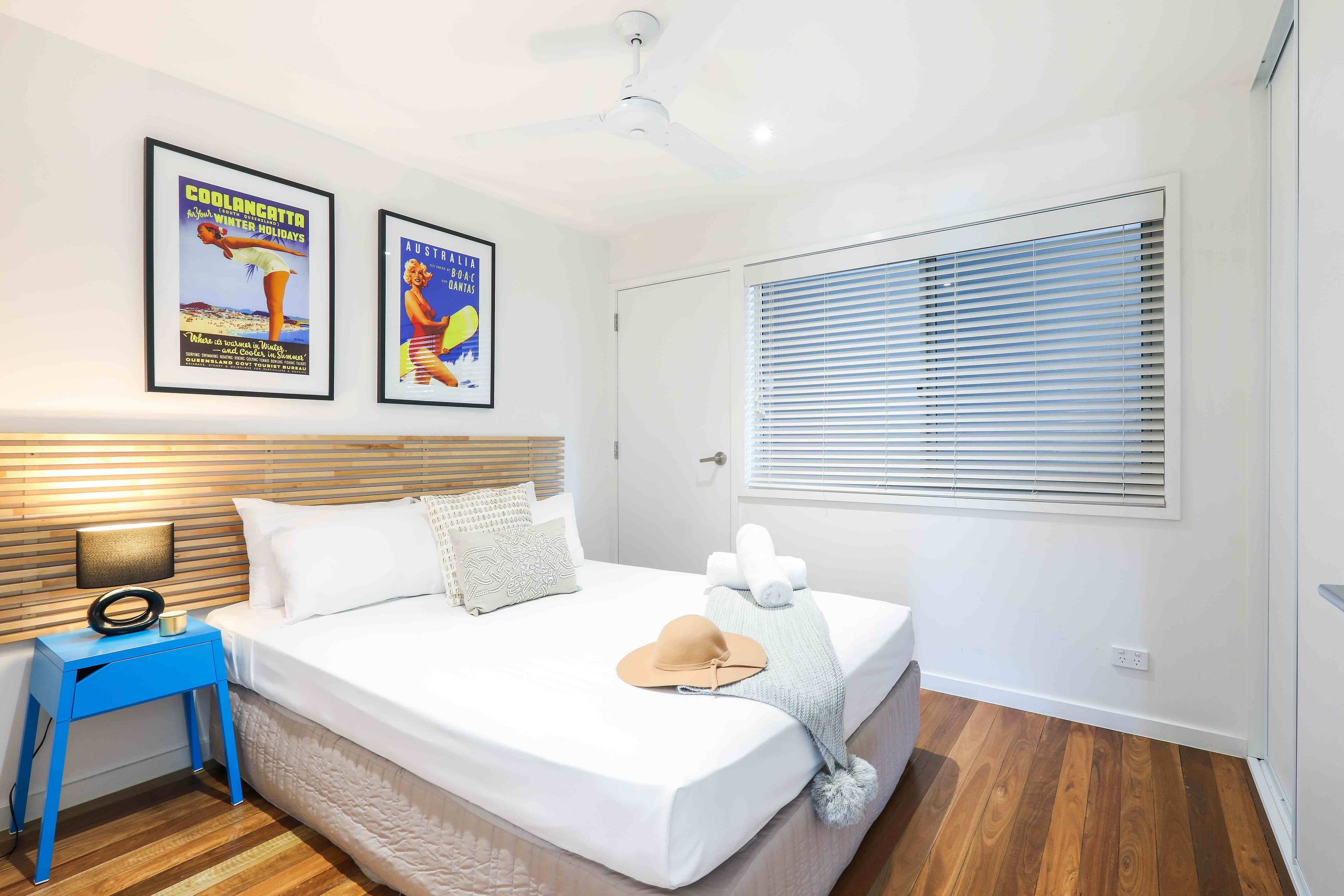 A neatly arranged bedroom with a bed, a nightstand, a ceiling fan, and two framed posters on the wall.