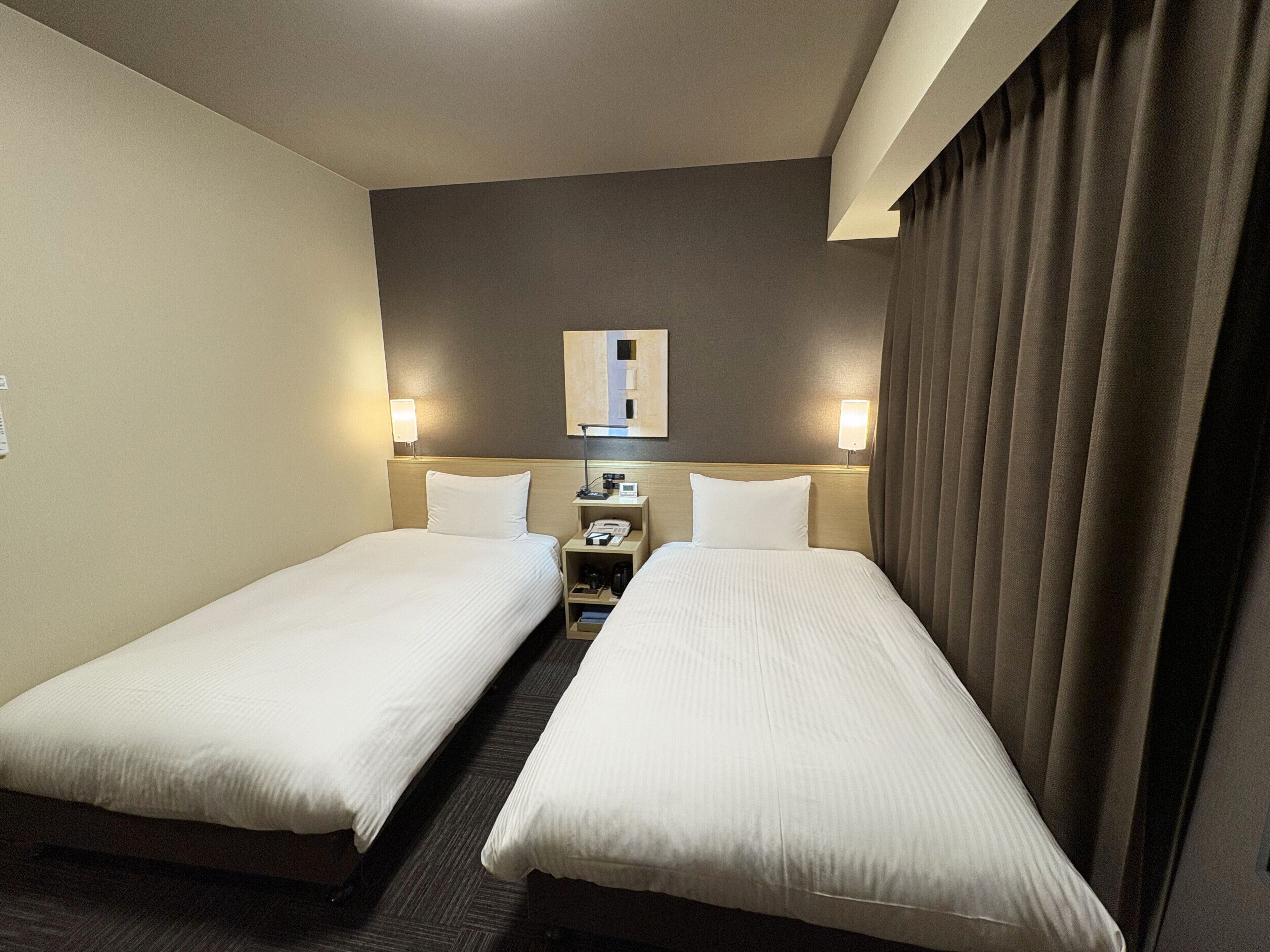 Quarto de hotel com duas camas de solteiro, mesa de cabeceira com abajur, espelho e janela com cortinas.