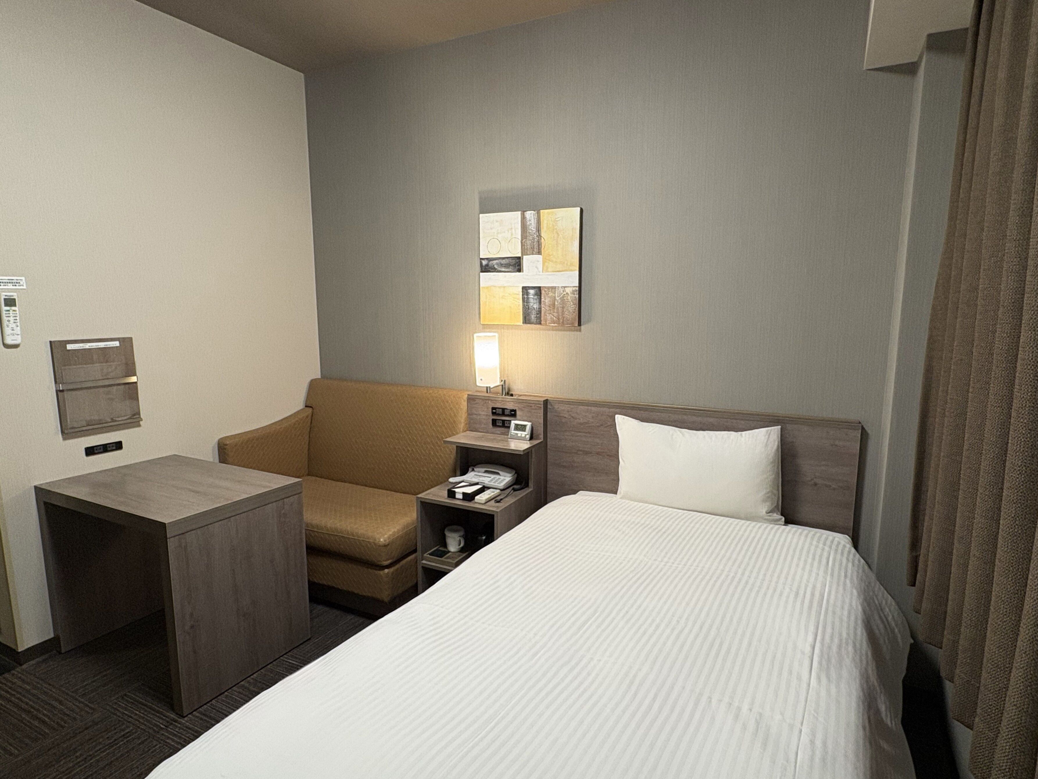 A hotel room with a bed, a desk, a chair, a nightstand with a lamp, a telephone, and a wall-mounted air conditioning unit.