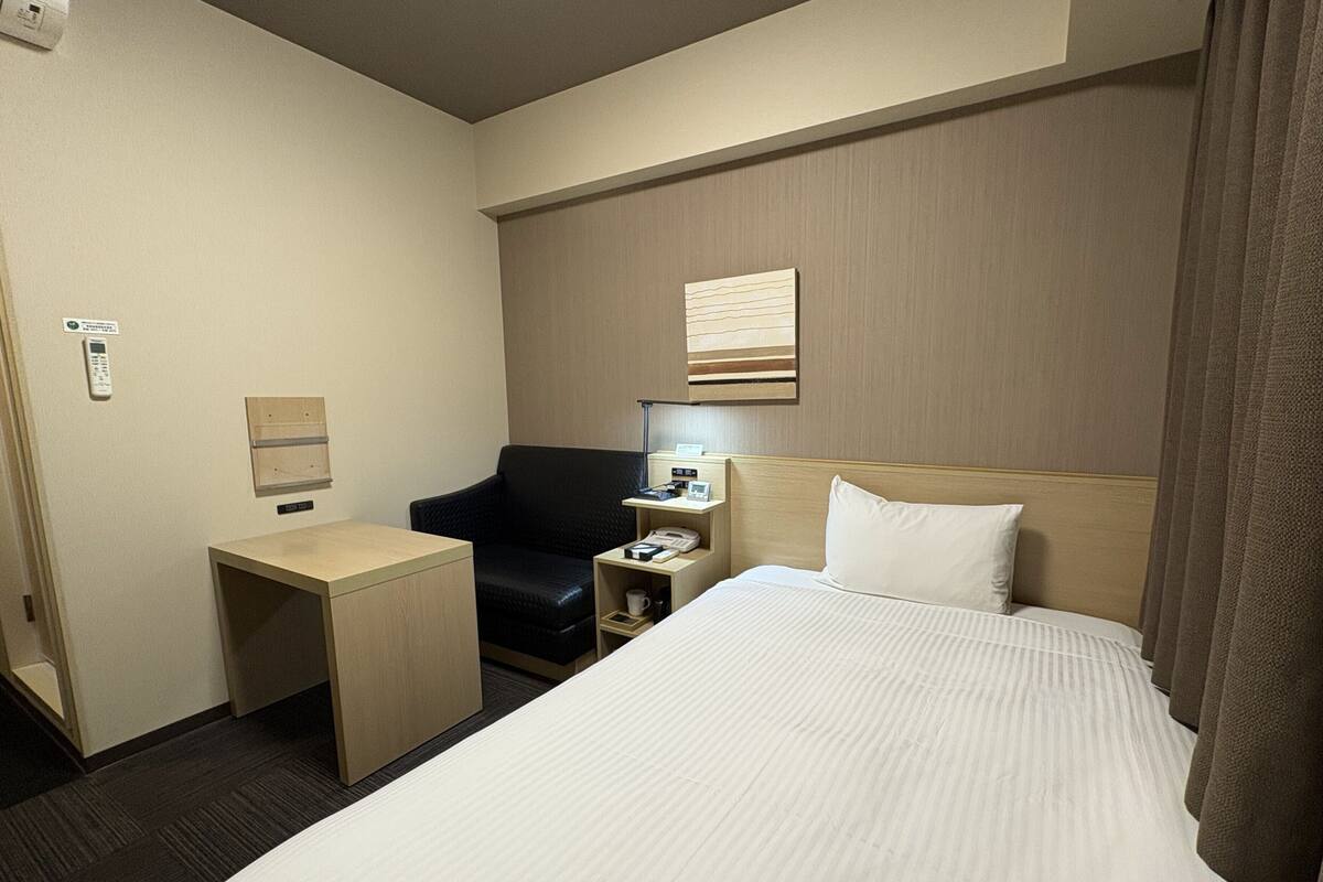 A hotel room with a bed, a desk, a chair, a nightstand, a lamp, and a wall-mounted artwork.