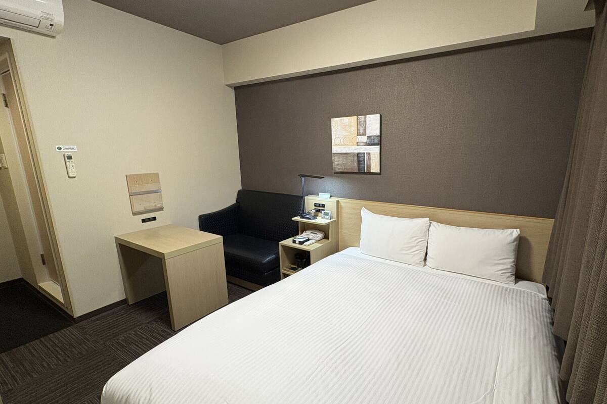 A hotel room with a bed, a desk, a chair, a nightstand, a telephone, and a wall-mounted artwork.