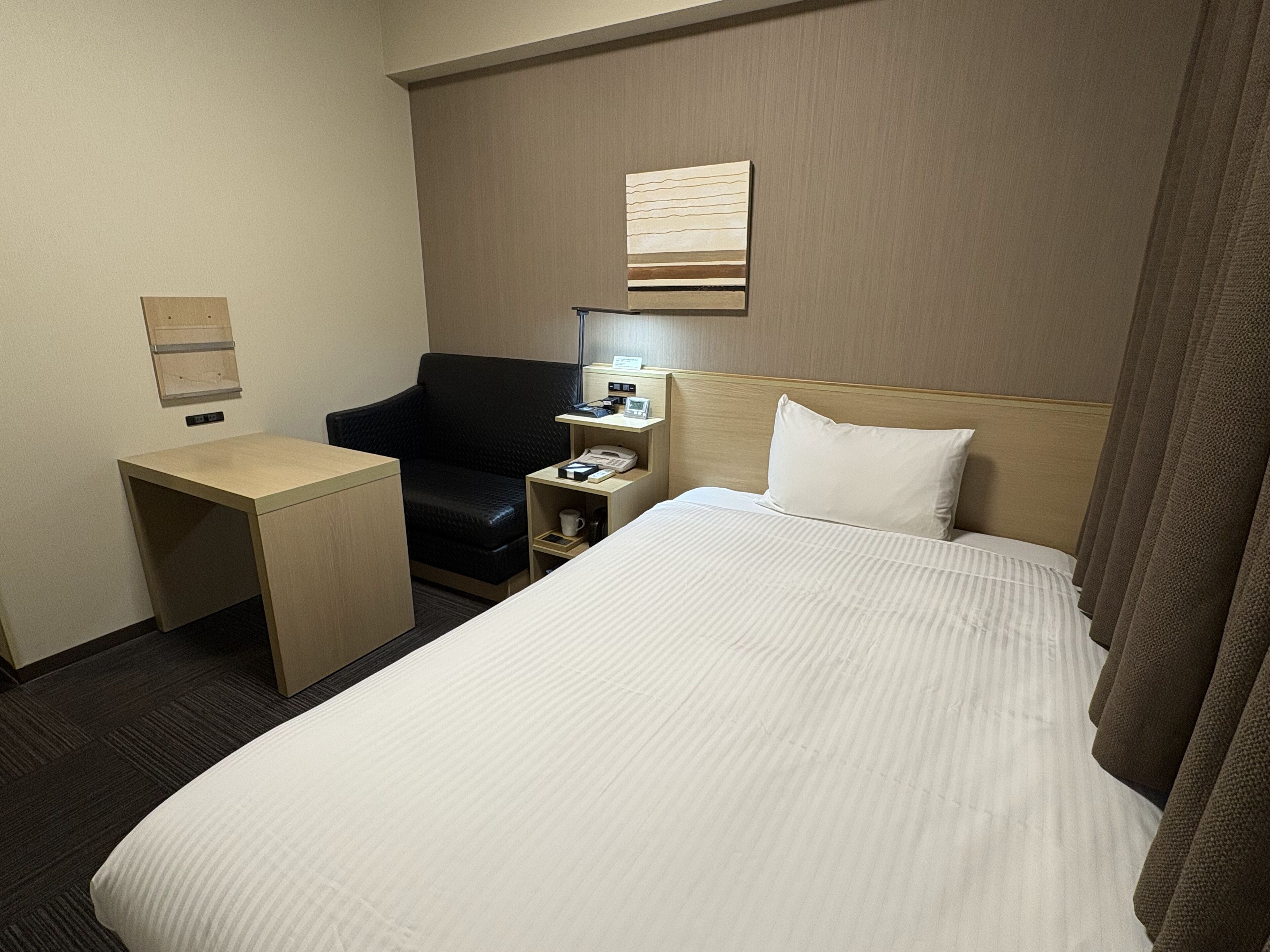 A hotel room with a bed, a desk, a chair, a nightstand, and a wall-mounted telephone.