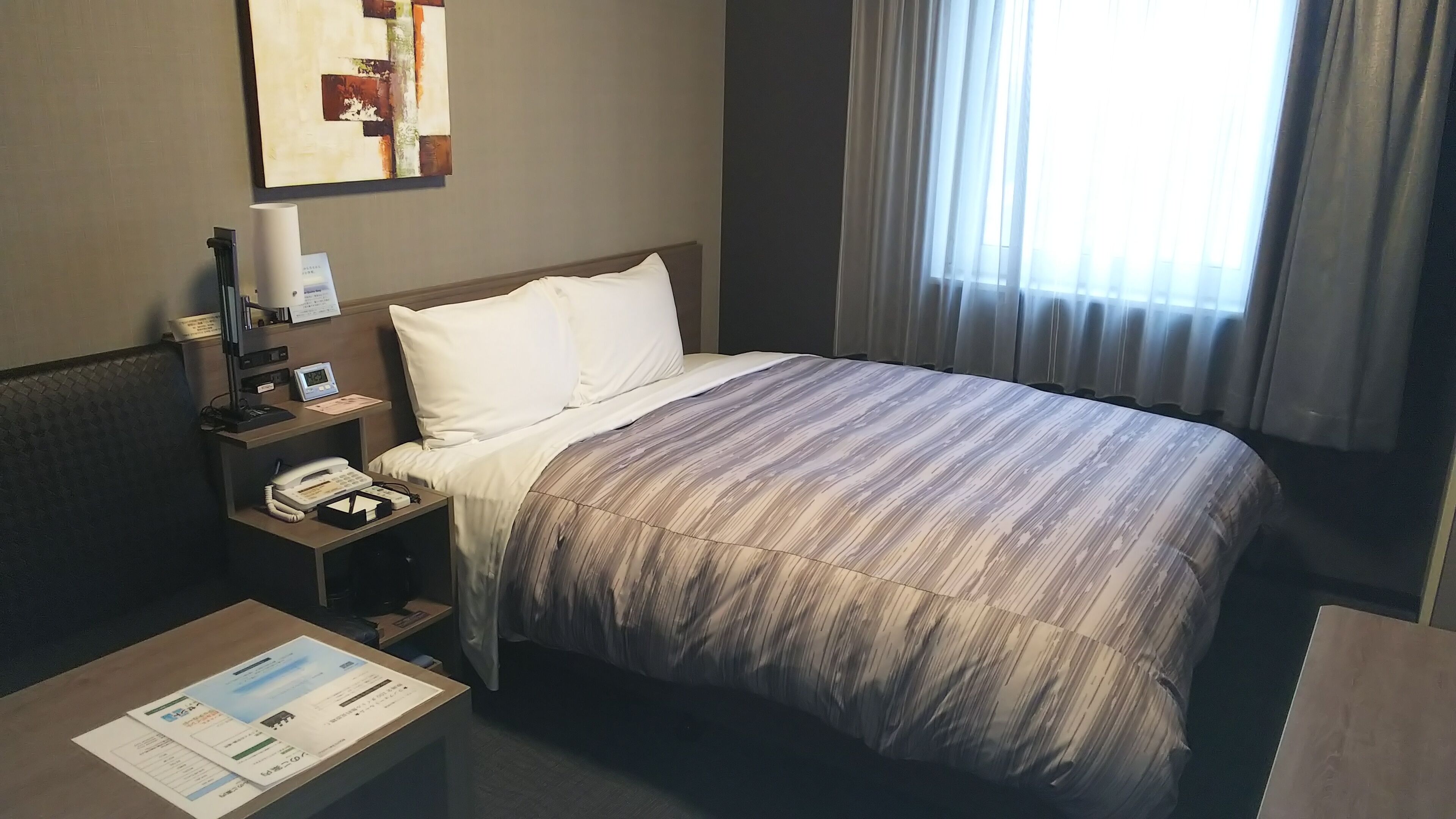 A hotel room with a bed, bedside table, lamp, clock, and a window with curtains.