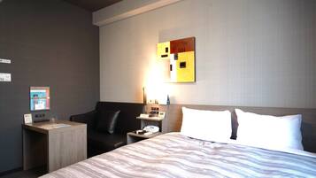 A hotel room with a large bed, a desk with a computer, a chair, a nightstand with a lamp, and a wall-mounted artwork.