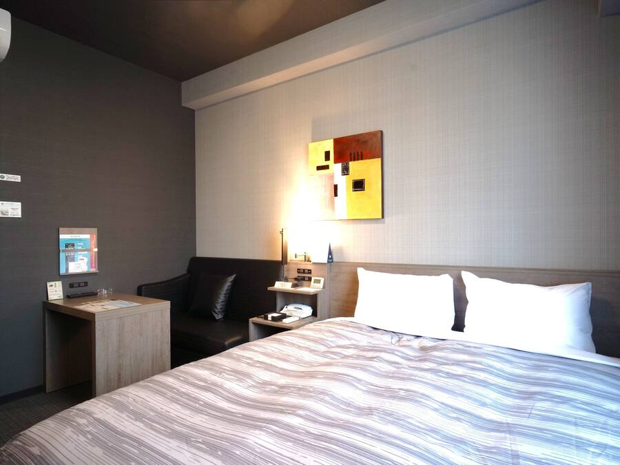 A hotel room with a large bed, a desk with a computer, a chair, a nightstand with a lamp, and a wall-mounted artwork.